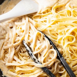 tongs grabbing cooked fettuccine and tossing in Alfredo sauce with spatula in homemade Alfredo sauce