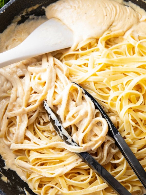 tongs grabbing cooked fettuccine and tossing in Alfredo sauce with spatula in homemade Alfredo sauce
