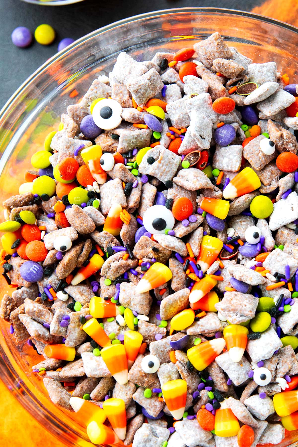 close up of this Halloween puppy chow recipe served in a bowl filled with candy corn, googly eyes, and purple and green sprinkles.