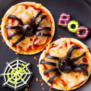 Halloween pizzas are adorably mini with melted cheese and black olive spiders, served on a party table with spooky decorations