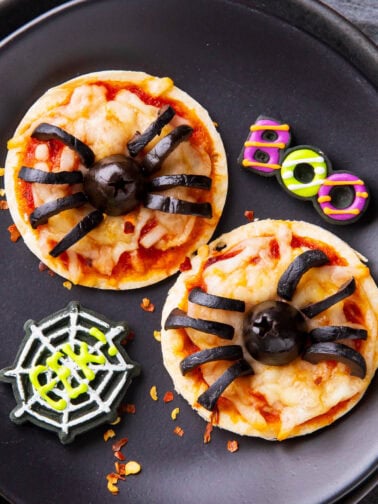 Halloween pizzas are adorably mini with melted cheese and black olive spiders, served on a party table with spooky decorations