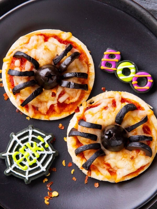 Halloween pizzas are adorably mini with melted cheese and black olive spiders, served on a party table with spooky decorations