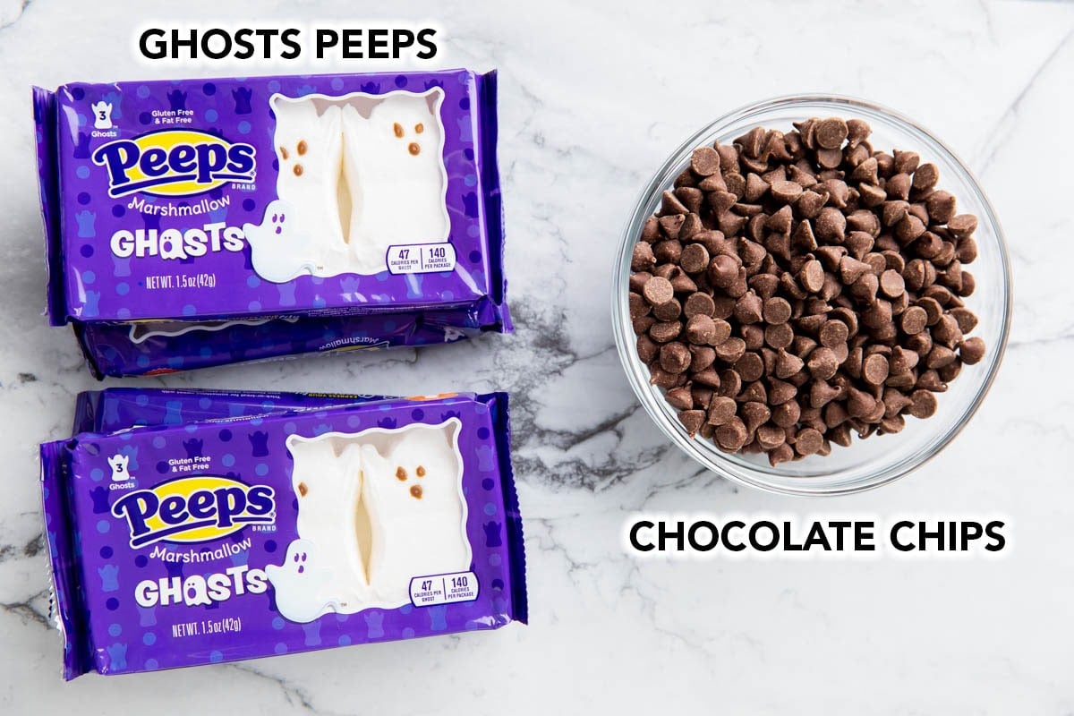 Halloween S'mores ingredients on a table including ghost Peeps marshmallows and chocolate chips.
