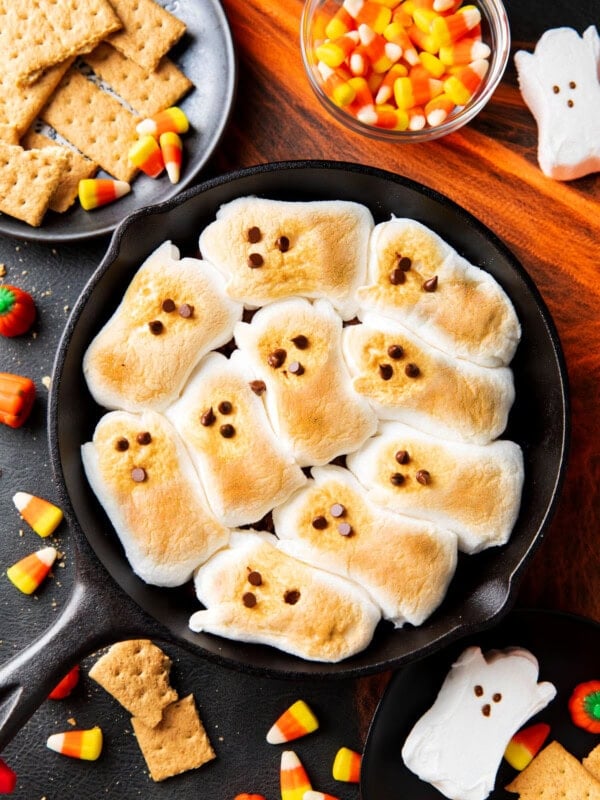 Halloween S'mores are topped with toasted ghost-shaped marshmallows serve on a Halloween party table with candy corn and graham crackers