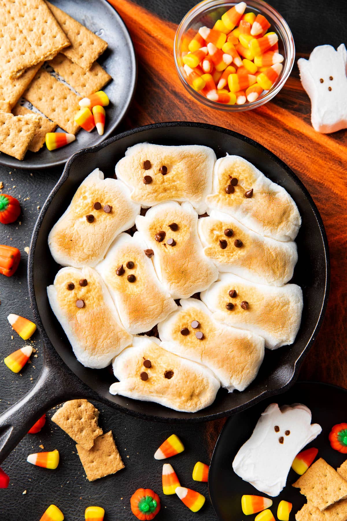Halloween S'mores are topped with toasted ghost-shaped marshmallows serve on a Halloween party table with candy corn and graham crackers.
