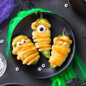 Mummy jalapeno poppers stuffed with cream cheese and wrapped in crescent roll dough is baked golden and served on a Halloween plate