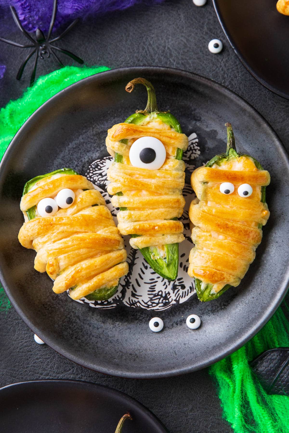 A Halloween table with appetizer plates of mummy jalapeno poppers wrapped in crescent roll dough and stuffed with seasoned cream cheese.
