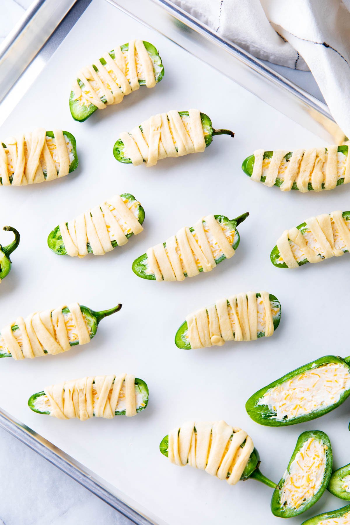 Cream cheese filled jalapeno popper mummies wrapped in crescent roll dough strips on a baking sheet before placing in oven.