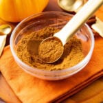 a measuring spoon scoops up pumpkin pie spice out of a spice bowl