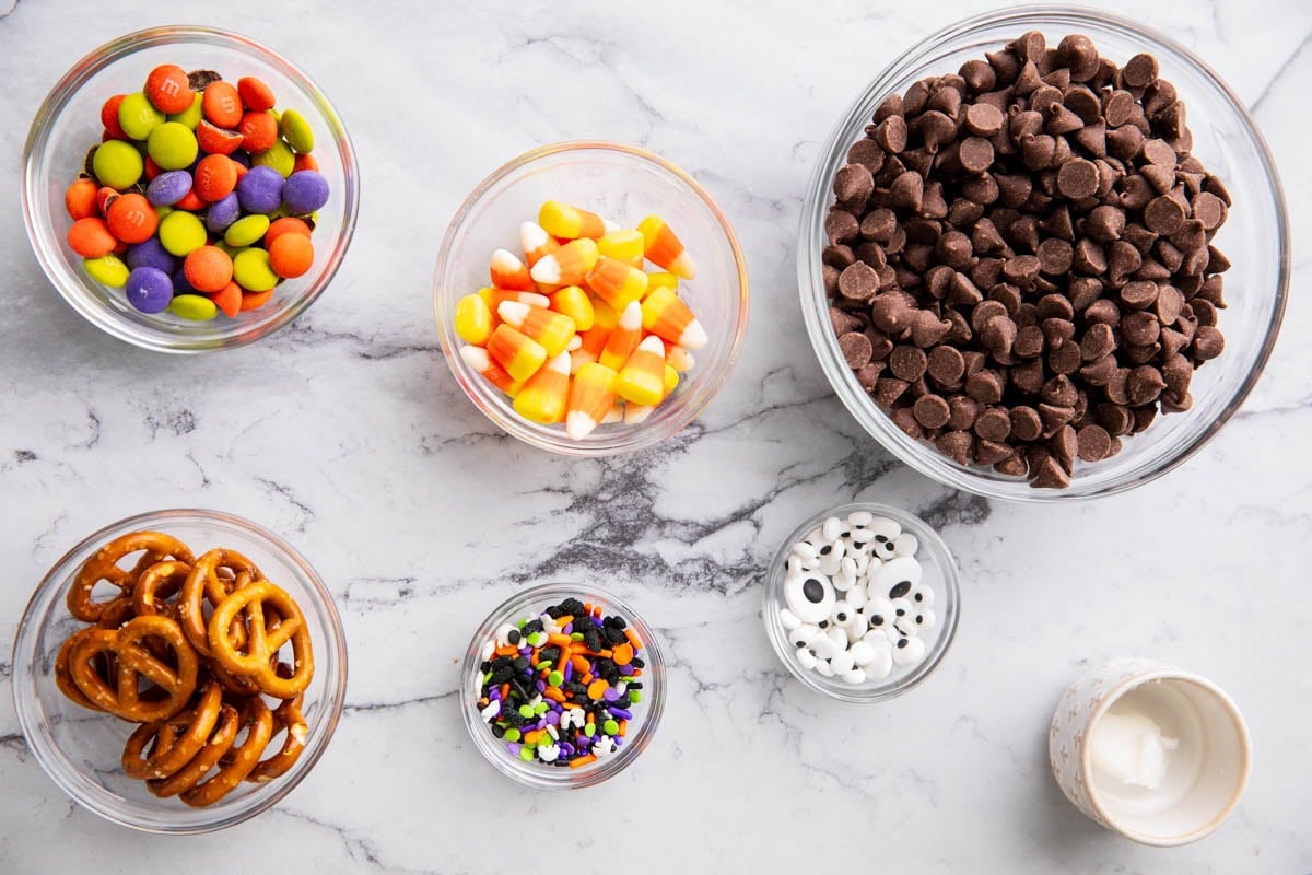 Halloween Bark ingredients measured into prep bowls including chocolate, candy corn, pretzels, googly eyes, Halloween sprinkles, and candies.