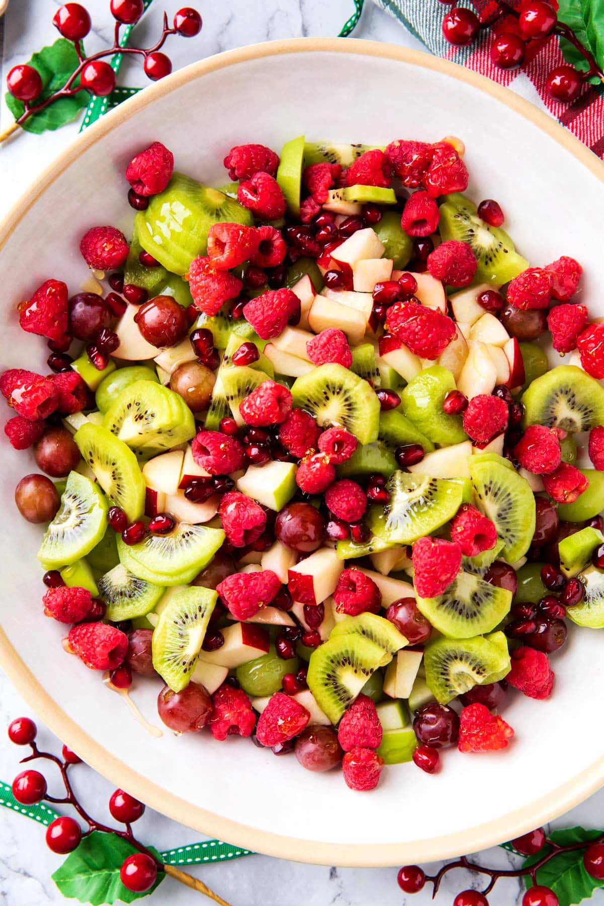 Serving this Christmas Fruit Salad recipe in a large bowl dotted with red raspberries, red and green fruit, and a honey lime dressing served with Christmas decor.