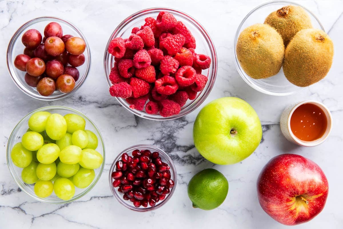 Christmas Fruit Salad ingredients laid out, including red and green grapes, kiwi fruit, red and green apples, pomegranate arils, lime juice, and honey.