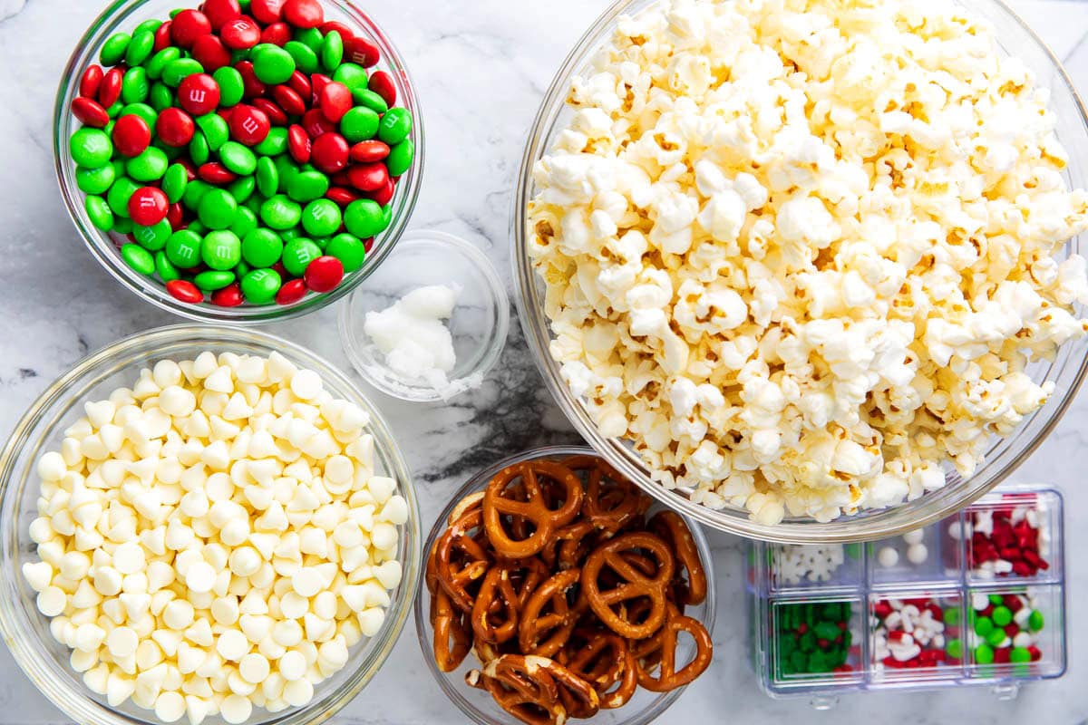 White chocolate popcorn ingredients measured into bowls including popcorn, white chocolate chips, M&M's, pretzels, and Christmas sprinkles.