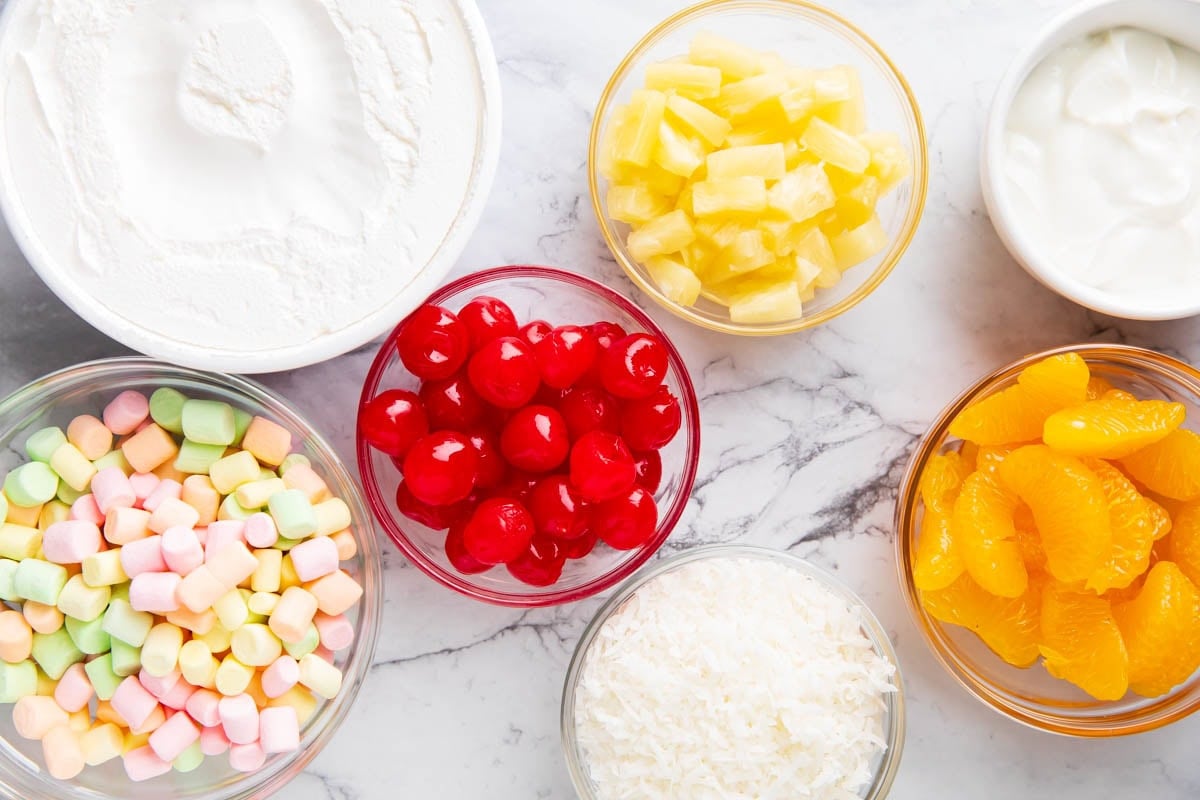 Ambrosa Fruit Salad ingredients include Cool Whip, vanilla yogurt, pineapple, cherries, mandarin oranges, fruit marshmallows, and sweetened coconut.
