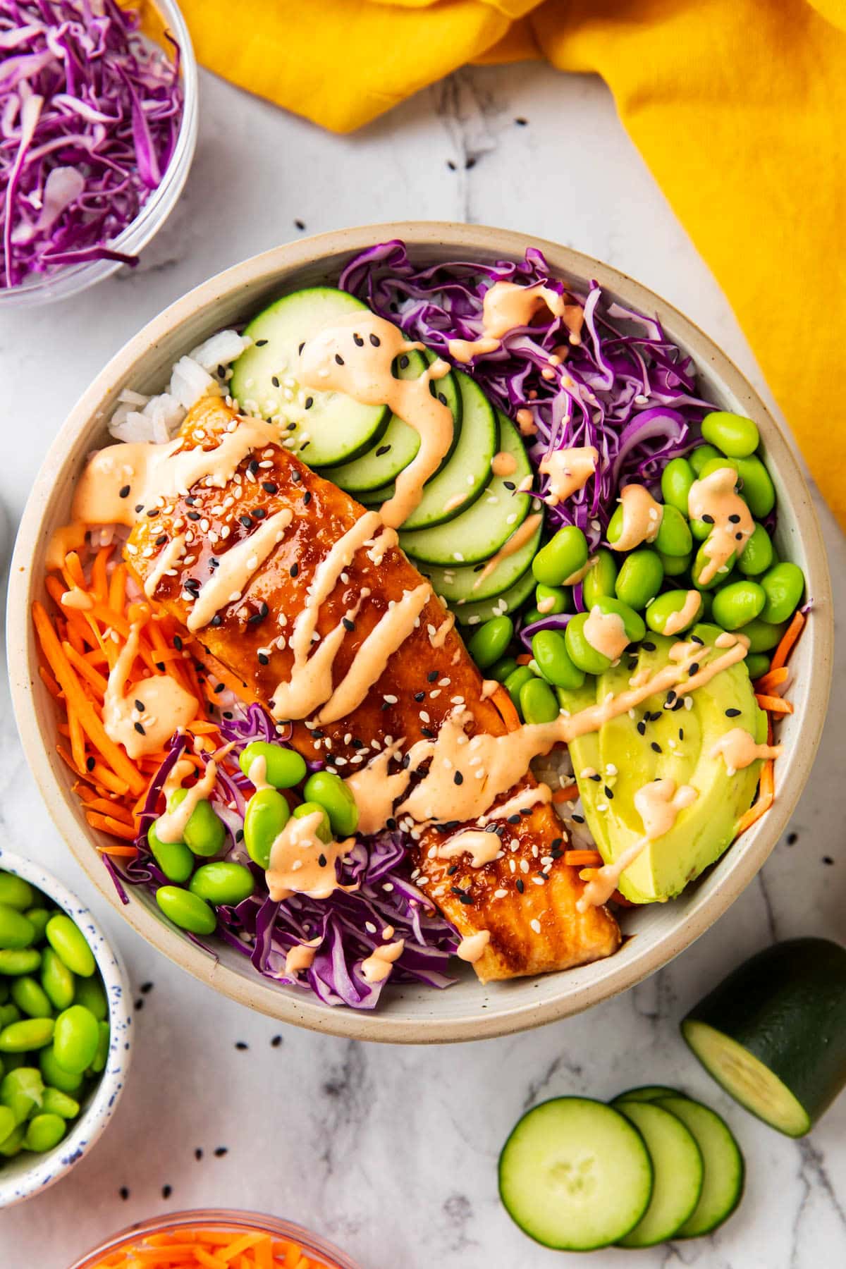 Salmon rice bowl is served with tender cooked salmon topped with sriracha mayo and served with rice, cucumber, avocado, cabbage, edamame, and carrots.