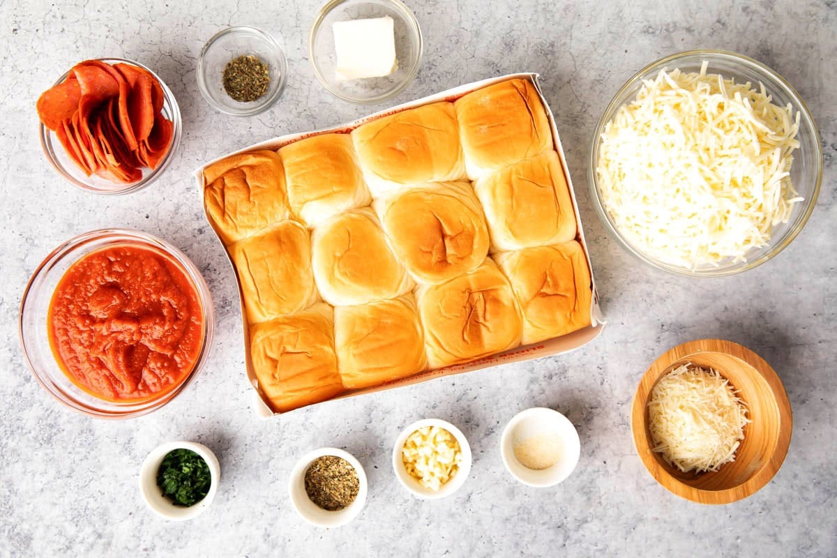 Pizza sliders ingredients in prep bowls, including pizza seasoning, Hawaiian rolls, cheese, pepperoni, butter, parmesan, and garlic.
