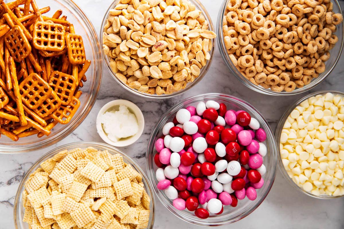 Valentines Chex Mix ingredients in prep bowls - Cheerios cereal, Chex cereal, peanuts, pretzel pieces, red, pink and white Valentine's M&M's, and white chocolate.
