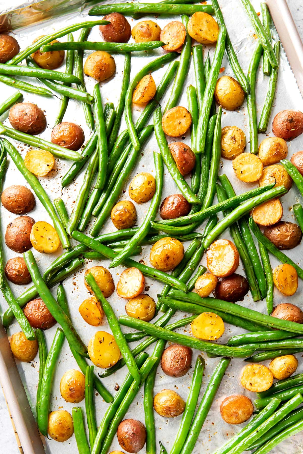 Roasting potatoes and green beans coated with olive oil and seasoning on a foil lined baking sheet.