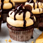 Chocolate cupcakes with peanut butter cups are topped with peanut butter frosting and chocolate drizzle