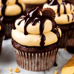 Chocolate cupcakes with peanut butter cups are topped with peanut butter frosting and chocolate drizzle