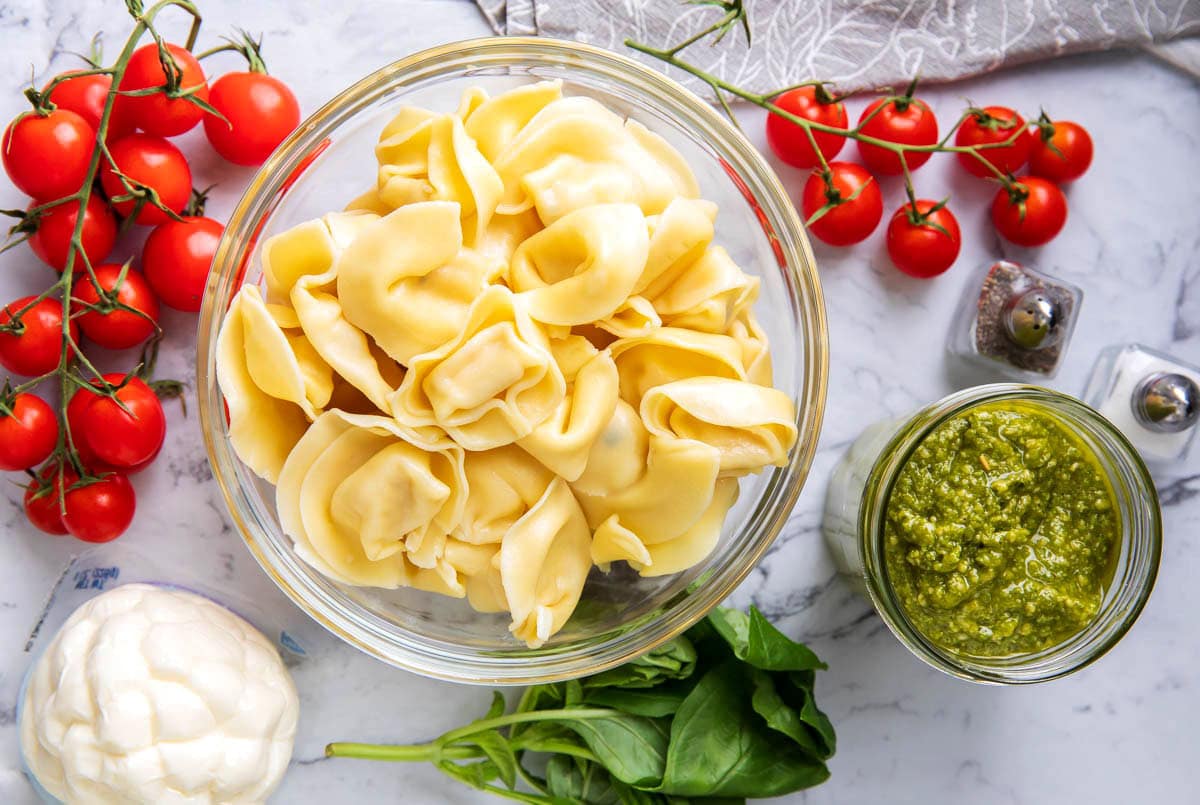 Easy basil tortellini pesto pasta salad ingredients - creamy basil pesto sauce, cheese tortellini, cherry tomatoes, and mozzarella pearls.