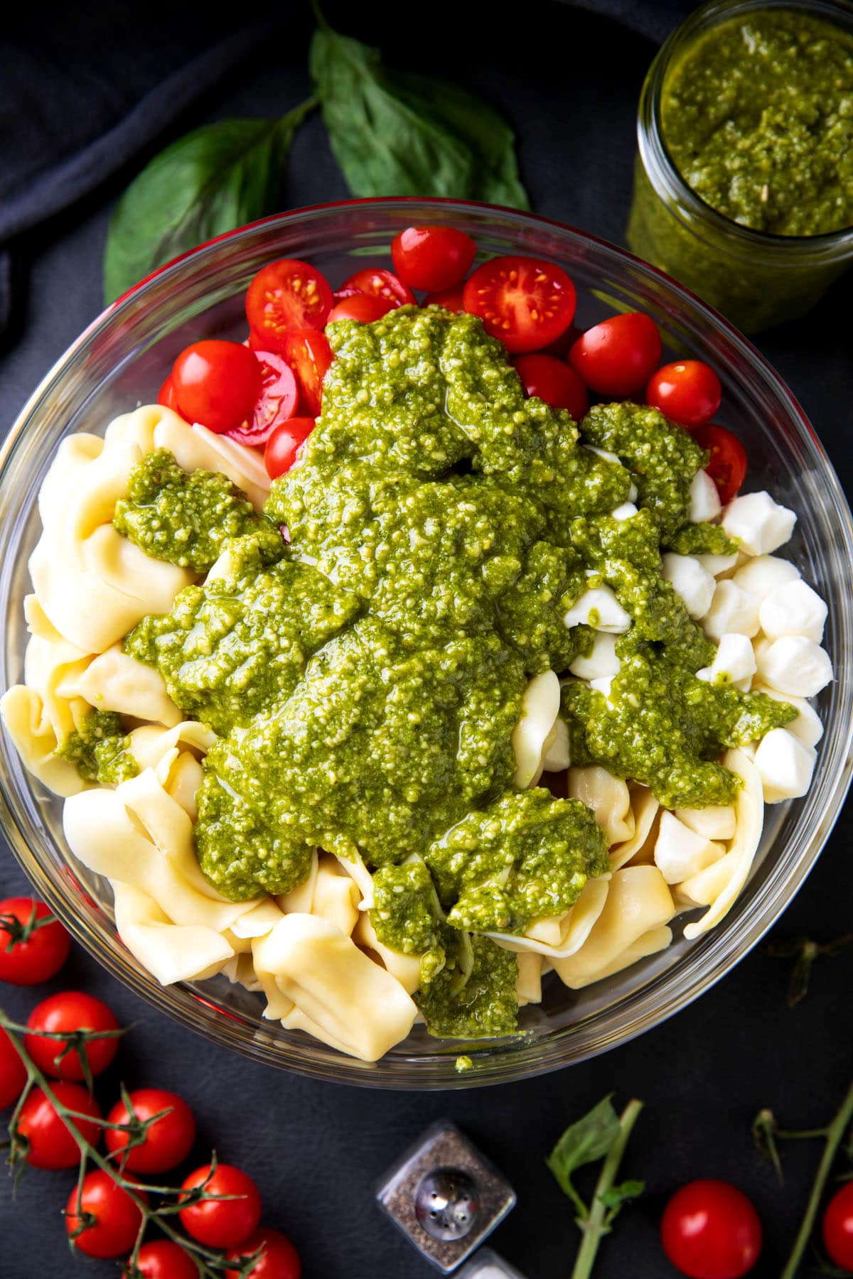 A photo showing how to make tortellini pesto pasta salad - basil pesto sauce poured over pasta, cheese, and tomatoes.