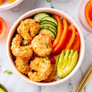 Bang bang shrimp rice bowls are topped with crispy, saucy bang bang shrimp, avocado, cucumbers, and bell peppers served over white rice in a bowl