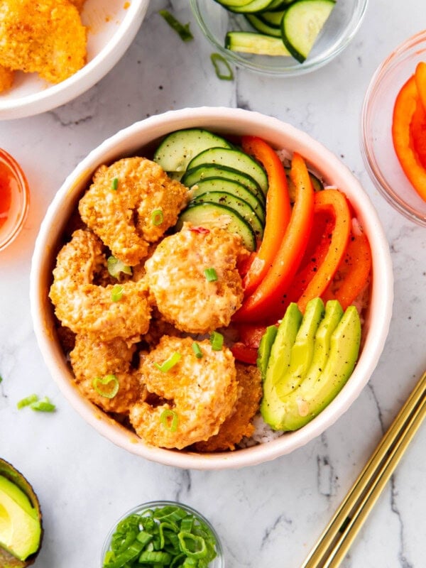 Bang bang shrimp rice bowls are topped with crispy, saucy bang bang shrimp, avocado, cucumbers, and bell peppers served over white rice in a bowl