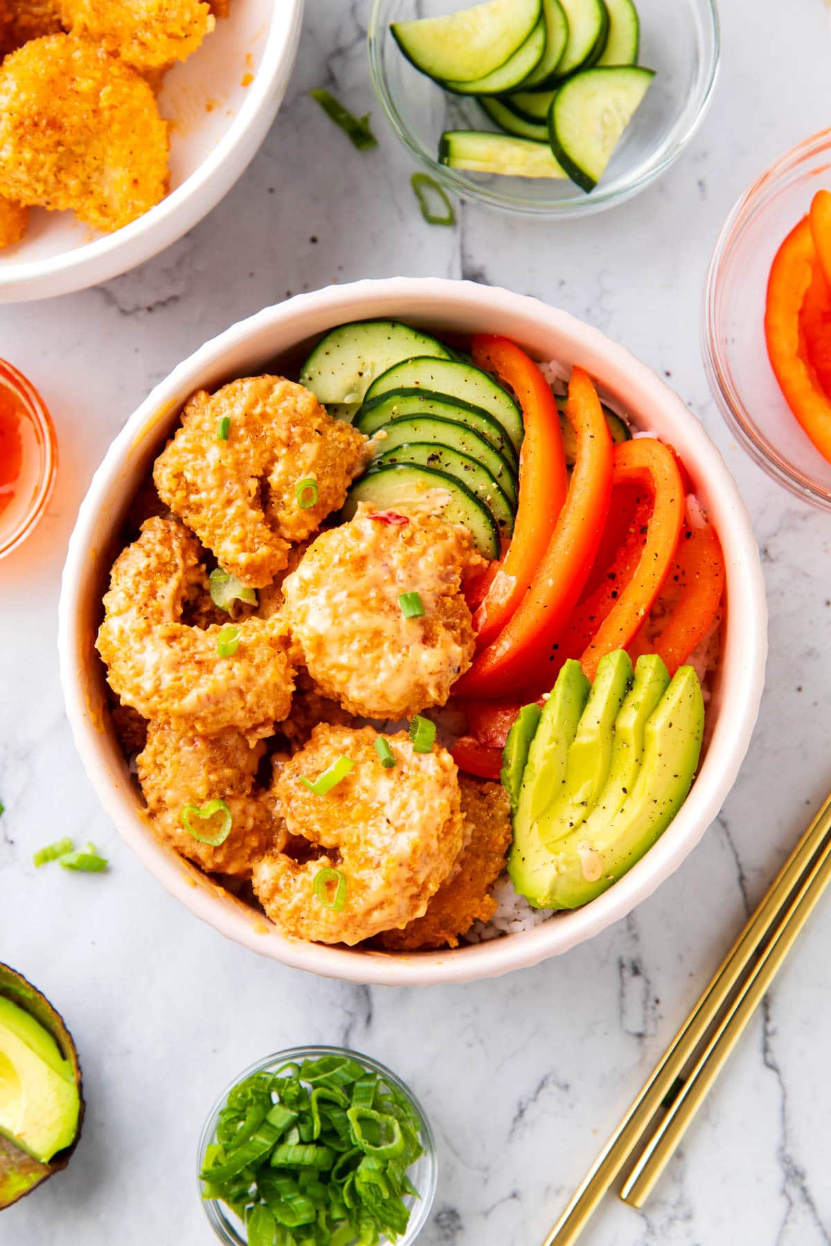 Bang bang shrimp rice bowls are topped with crispy, saucy bang bang shrimp, avocado, cucumbers, and bell peppers served over white rice in a bowl.