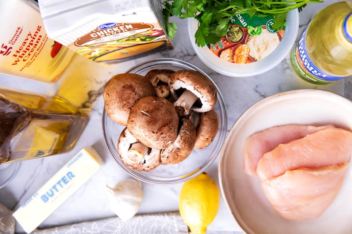 Creamy mushroom chicken ingredients on a table including mushrooms, chicken broth, chicken breast, heavy cream, olive oil, butter, Parmesan, lemon, garlic and dry white wine.