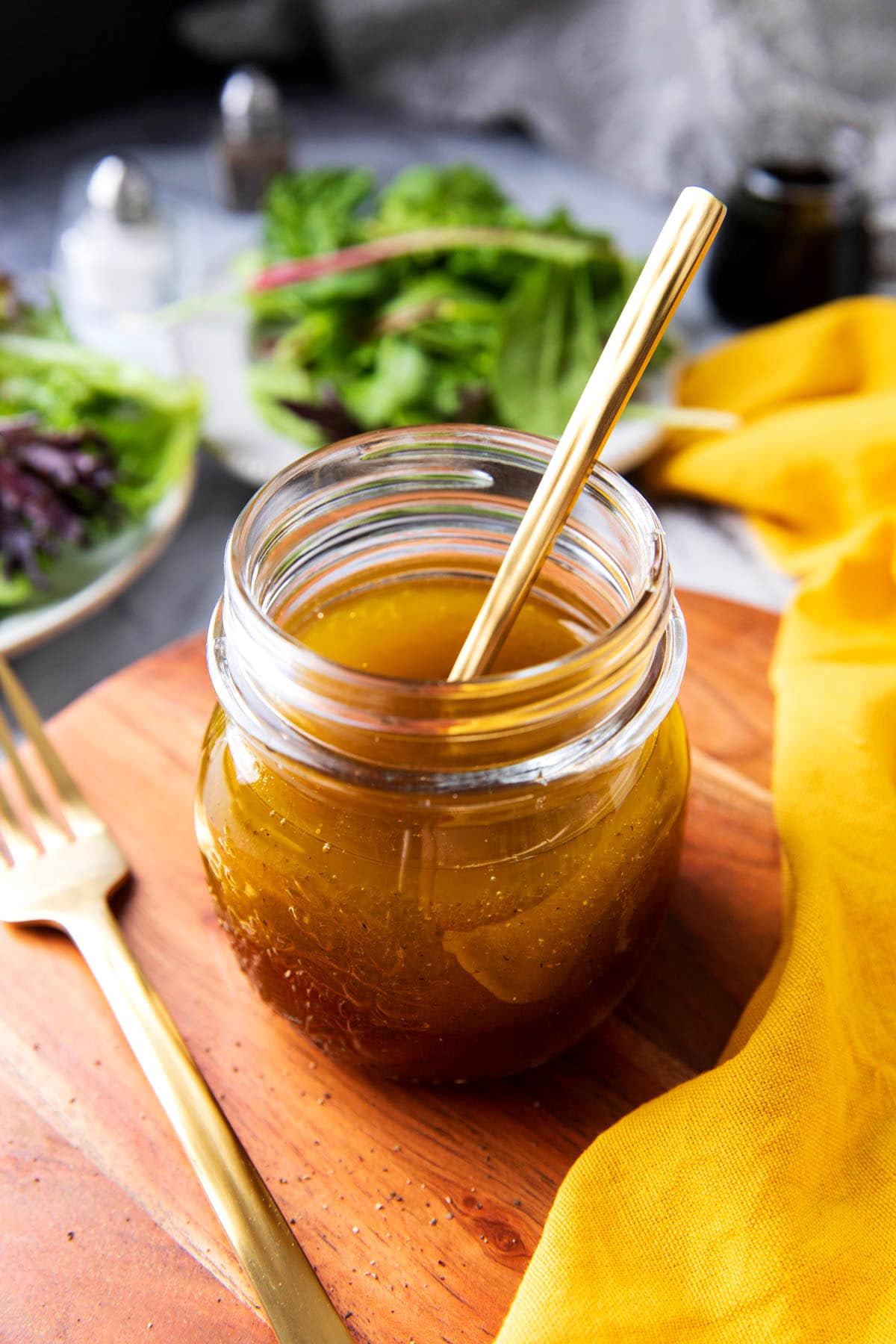 A bottle of Maple Vinaigrette sits with a spoon in it near two plates of salad.