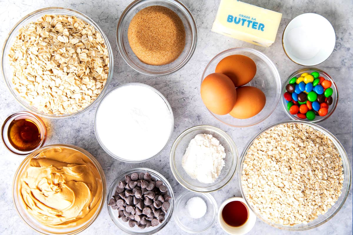 Monster Cookie Bars ingredients in prep bowls including oats, flour, baking soda, peanut butter, maple syrup, butter, vanilla, chocolate chips, eggs, brown sugar, and M&M's candies.