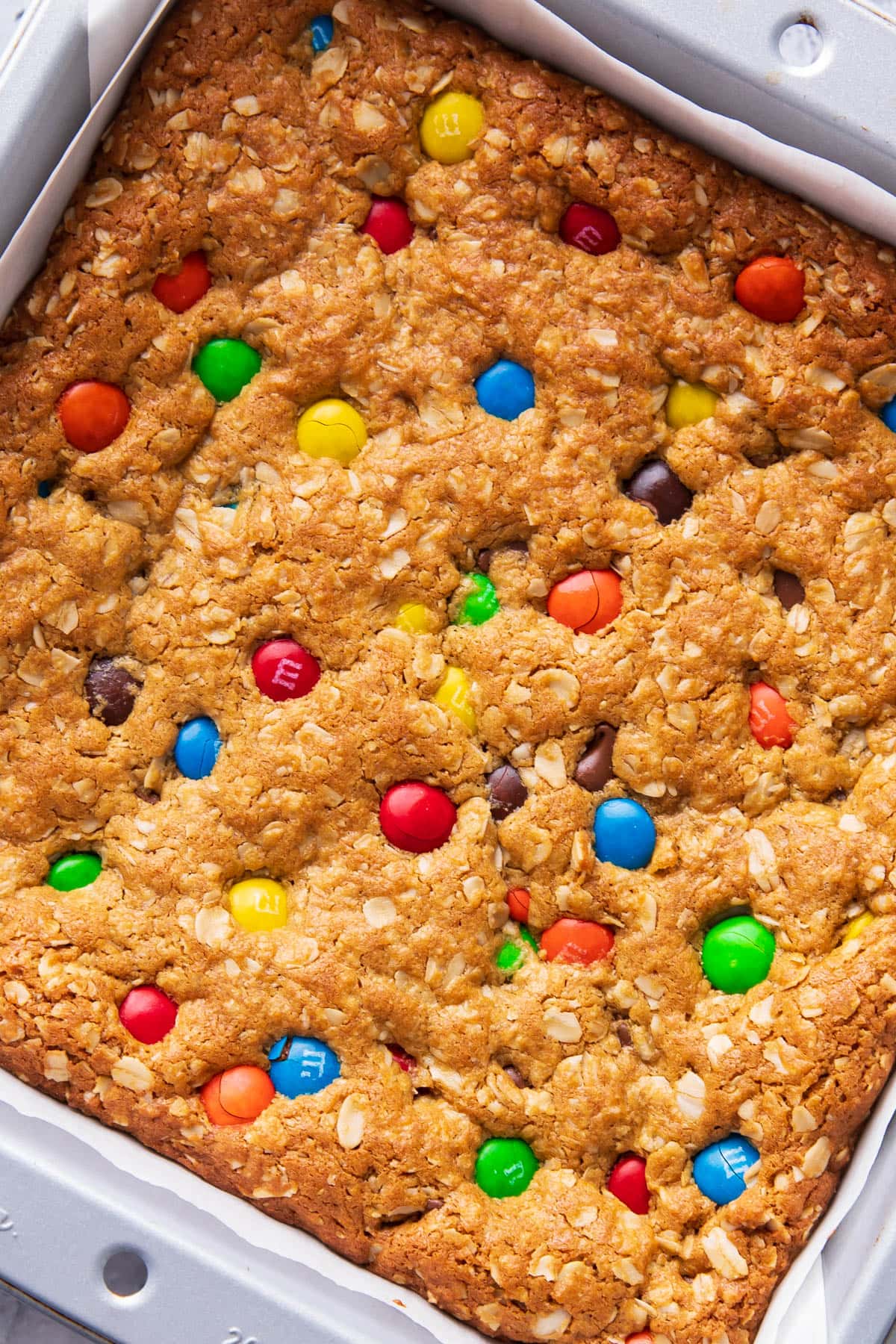 Close up of baked goods with M&M's in a lined pan to show golden, chewy texture with crisp edges topped with loads of oats, chocolate chips, and candies.