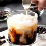 Close up of someone pouring milk from a jigger over coffee liqueur to make a White Russian