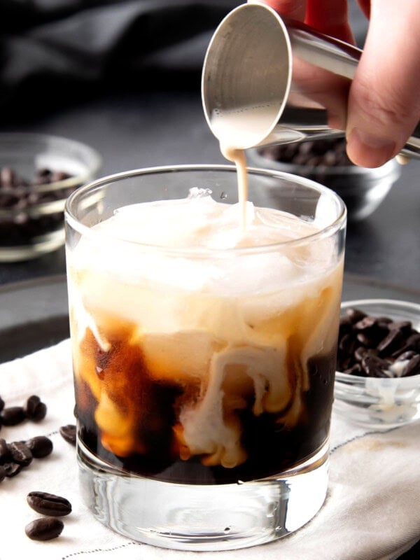 Close up of someone pouring milk from a jigger over coffee liqueur to make a White Russian