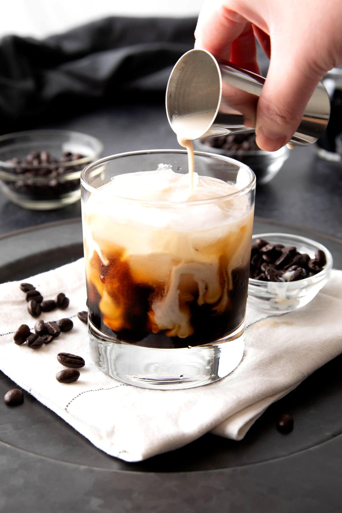 A photo showing How to Make a White Russian with Milk - pouring heavy cream over Kahlua and ice in a glass.